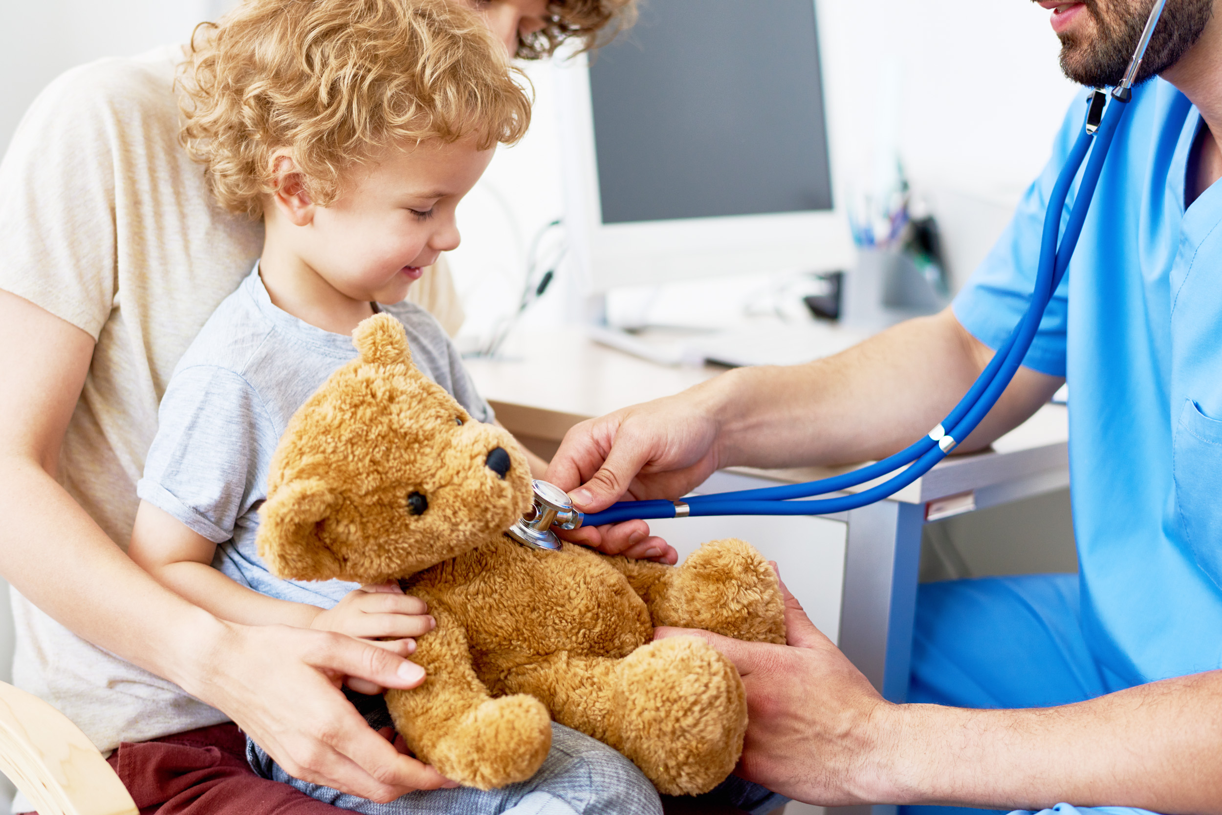 Portrait of adorable curly child sitting on mothers lap in doctors office holding teddy bear toy, with pediatrician listening to heartbeat using stethoscope