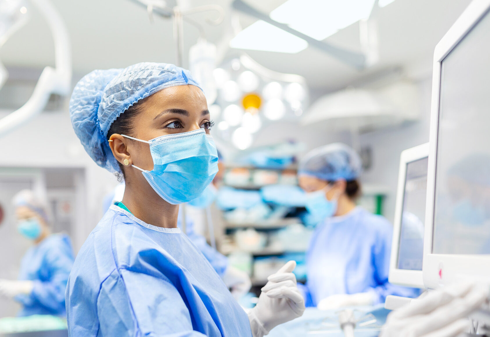 Anesthetist Working In Operating Theatre Wearing Protecive Gear checking monitors while sedating patient before surgical procedure in hospital