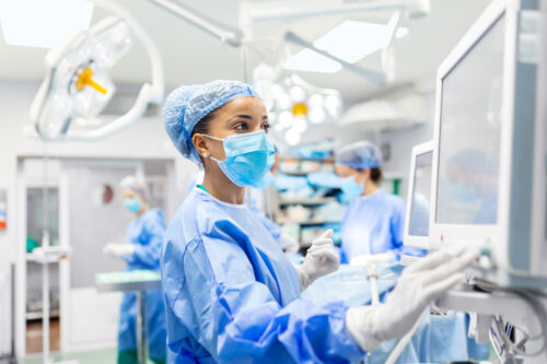 Anesthetist Working In Operating Theatre Wearing Protecive Gear checking monitors while sedating patient before surgical procedure in hospital