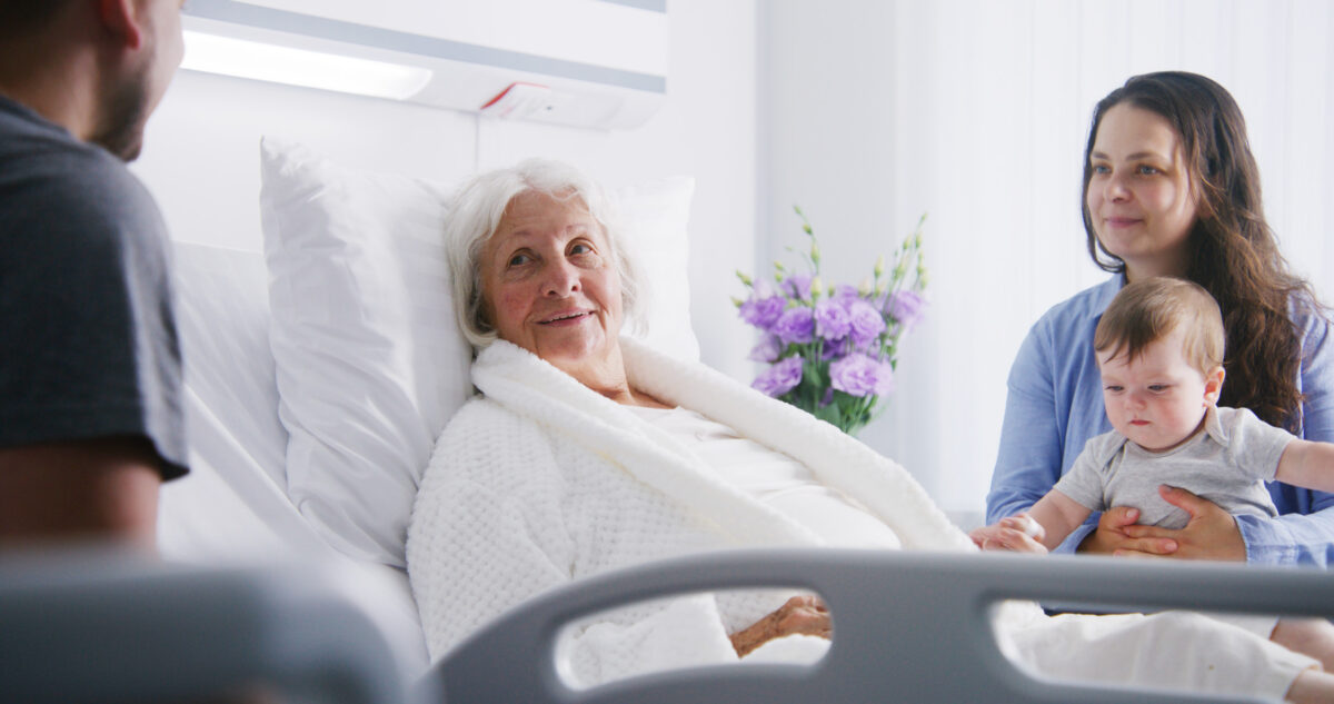Female old patient lies in bed in bright hospital ward, spends time with relatives. Loving family members visit elderly woman recovering after successful surgery. Modern medical facility or clinic.